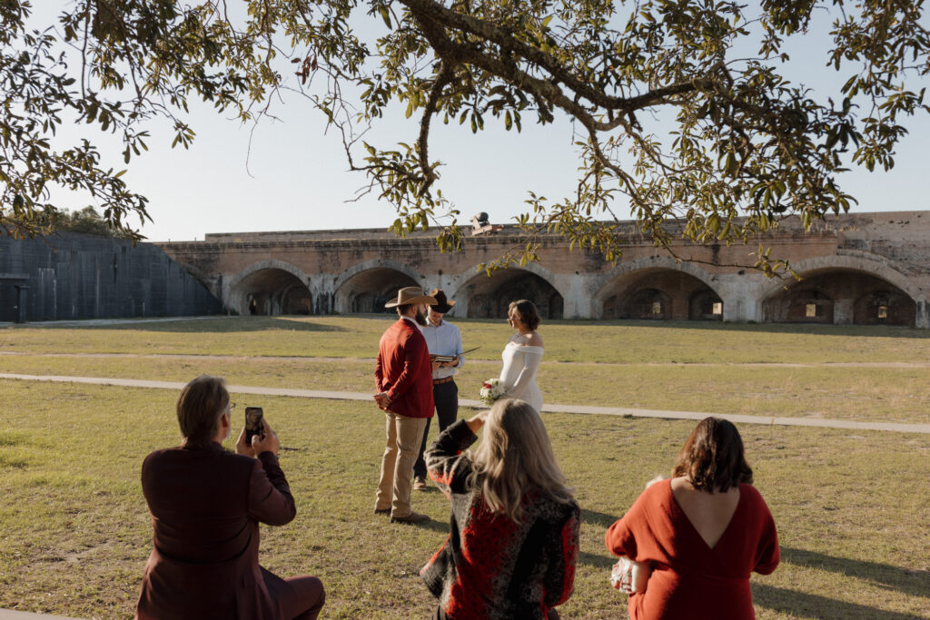 Fort Pickens elopement ceremony under a tree with benches, surrounded by family and sunlight